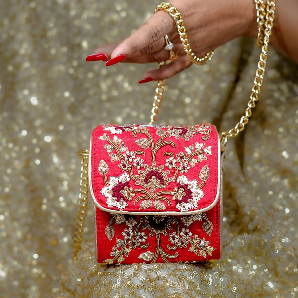 Naina red box bag held by its gold chain against a gold sequin lehenga